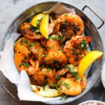 Spicy garlic shrimp garnished with herbs and lemon wedges in a metal bowl.