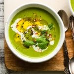 A bowl of vibrant green soup garnished with cream, basil, and spices.