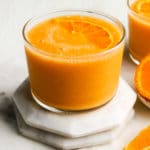 A glass of orange smoothie with fresh orange slices on a coaster.