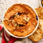 Bowl of muhammara dip topped with chopped walnuts, surrounded by crackers and vegetables.