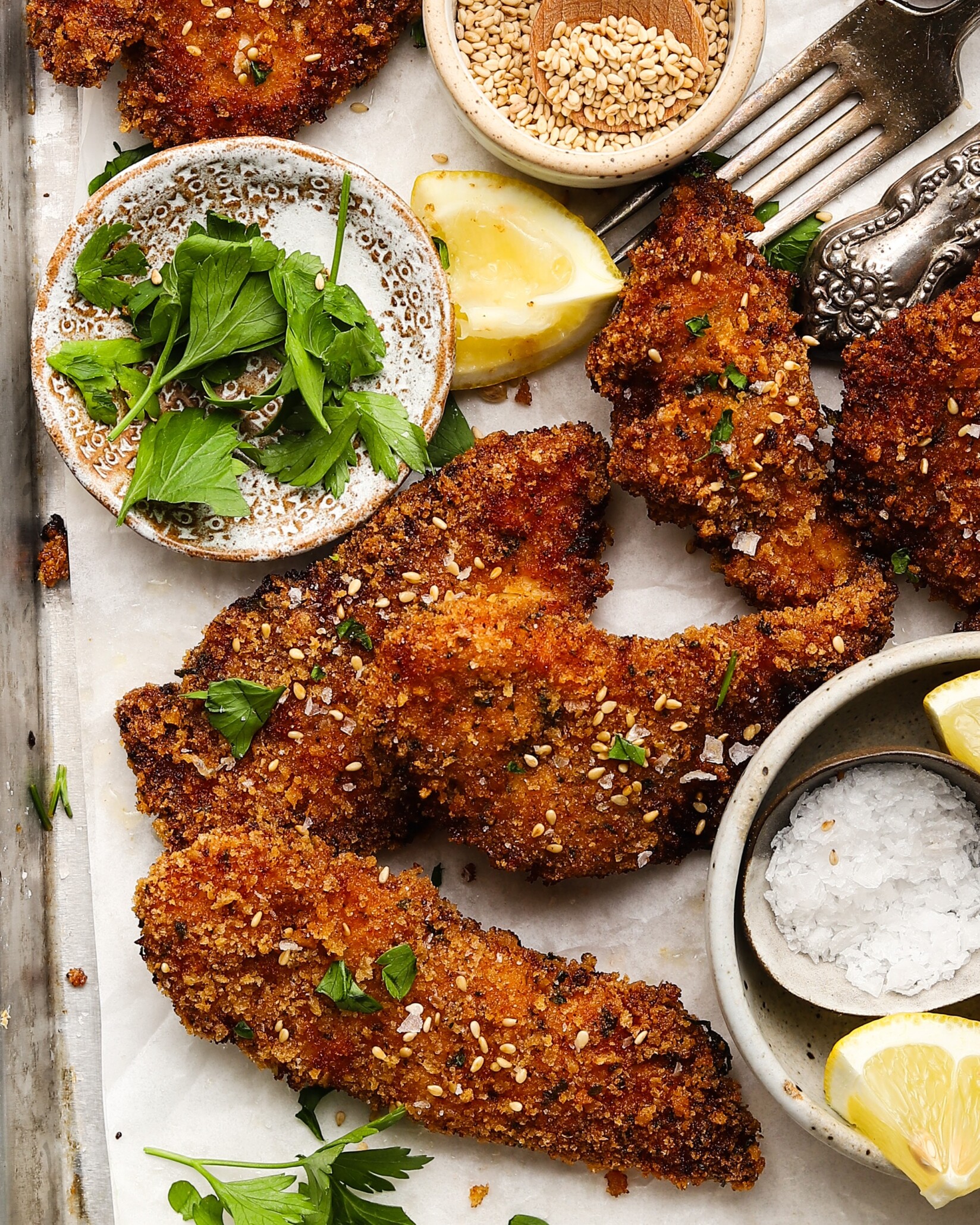 Crispy chicken schnitzel with lemon wedges, parsley, sesame seeds, and salt.