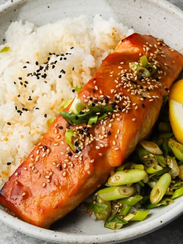 A dish of glazed salmon, sesame seeds, rice, scallions, and lemon wedges.