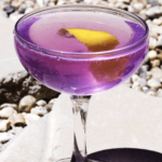 Purple cocktail in a coupe glass with a lemon twist garnish.