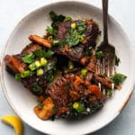 Braised short ribs garnished with green onions and herbs in a white bowl.