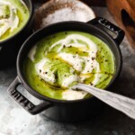 Green broccoli soup with crème fraîche, black pepper, and a silver spoon in bowl.