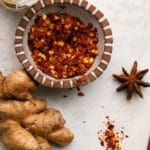 Bowl of chili flakes with ginger, garlic, and star anise on a light surface.