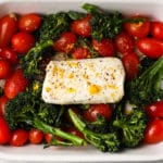 Baked goat cheese with cherry tomatoes and broccolini in a white baking dish.