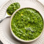 Bowl of vibrant green herb pesto with a spoon on a white plate.
