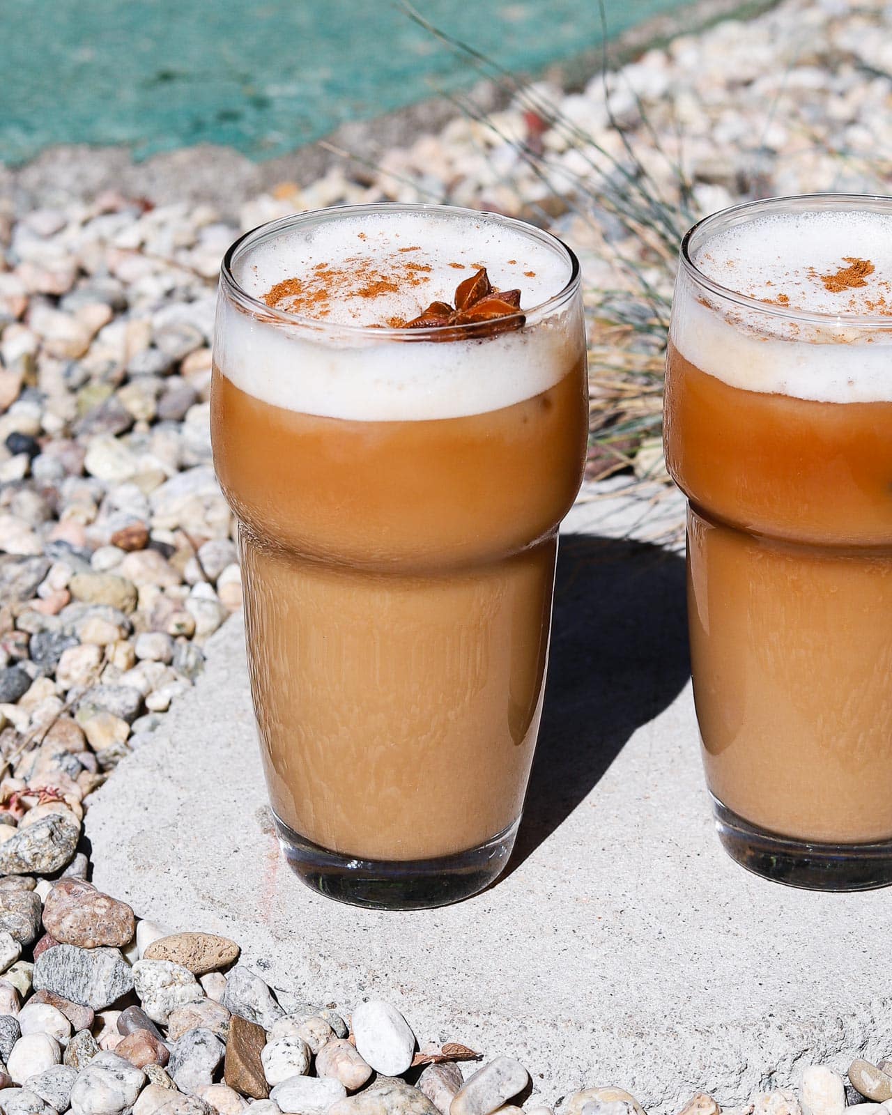 Two iced chai lattes topped with cinnamon and star anise, outdoors on gravel.