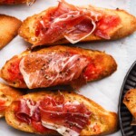 Toasted bread topped with tomato and cured ham slices, garnished with black pepper.