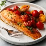 Seared salmon with burst cherry tomatoes garnished with herbs on a white plate.