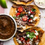 Three braised lamb tacos with tomato salsa, cilantro, and cheese beside a salsa bowl.