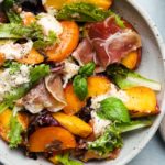 Salad with peaches, burrata, prosciutto, fresh greens, and basil leaves on a plate.