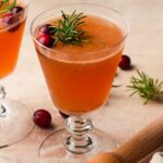 Refreshing cranberry drink with rosemary garnish in a glass, surrounded by fresh cranberries.