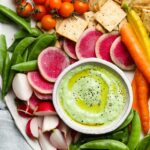 Green yogurt dip with fresh vegetables, crackers, tomatoes, and radish slices on a plate.