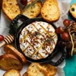 Baked camembert topped with honey and chili flakes, surrounded by toasted bread and grapes.