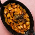 Roasted garlic cloves with rosemary in a black baking dish on a pink surface.