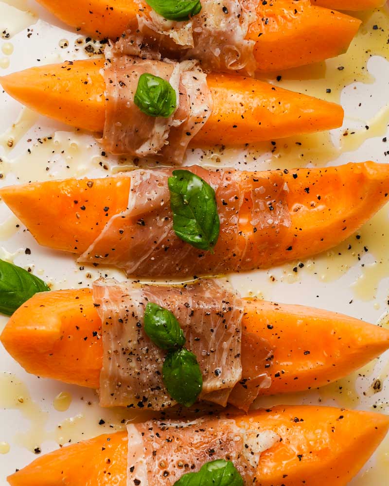 Sliced melon wrapped in prosciutto, garnished with basil and cracked black pepper.