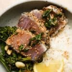 Seared albacore tuna with sesame seeds served over rice and greens, garnished with lemon.