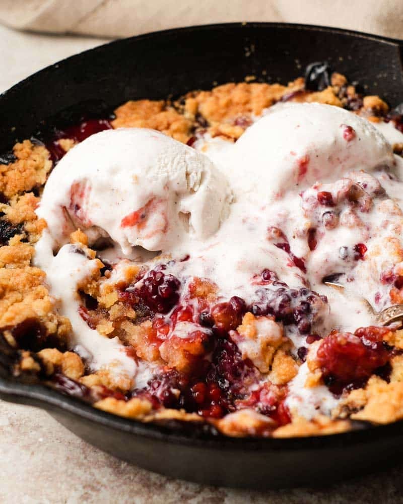 Mixed berry cobbler topped with melting vanilla ice cream in a cast iron skillet.