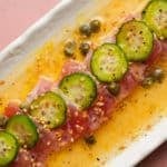 Tuna crudo with cucumber slices, capers, and sesame in citrus vinaigrette on plate.