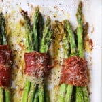 Prosciutto-wrapped asparagus topped with melted cheese and seasoning on a baking sheet.