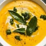 Creamy roasted pumpkin soup with pesto and garnished sage leaves in a white bowl.