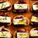 Toasted bread topped with cranberry sauce, brie, rosemary, and pepper on a wooden board.