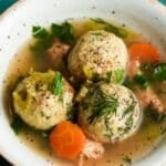 Matzo ball soup with carrots, herbs, and black pepper in a white bowl.