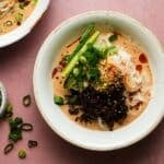 Bowl of sesame noodles with mushrooms, scallions, and cucumbers on pink background.