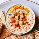 Bowl of homemade hummus topped with chickpeas, paprika, and olive oil, surrounded by pita.