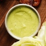 Bowl of creamy green basil mayo on a wooden surface near lettuce.