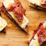Toasted bread topped with prosciutto, creamy brie, and jam on a wooden board.