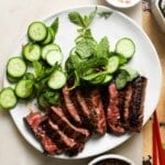 Grilled Thai steak with cucumber slices and mint leaves on a white plate.
