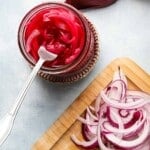 Jar of pickled red onions with sliced onions on a wooden board.