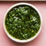 Bowl of green chimichurri sauce on a pink surface.