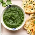 Green cilantro chutney in a bowl with naan bread and fresh cilantro on the side.