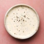 A bowl of creamy ranch dressing with herbs and pepper on a pink background.