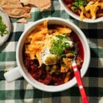 A bowl of chili topped with shredded cheese, pickled jalapeños, and green onions with an orange spoon.