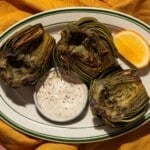 Close-up of grilled artichokes with aioli and a fresh slice of lemon on the plate.