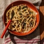 Red bowl filled with pasta, capers, cheese, and fresh herb garnish.