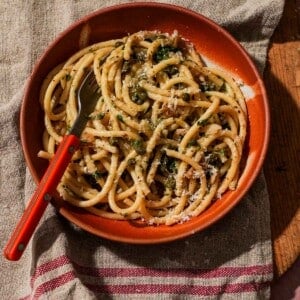 Red bowl filled with pasta, capers, cheese, and fresh herb garnish.