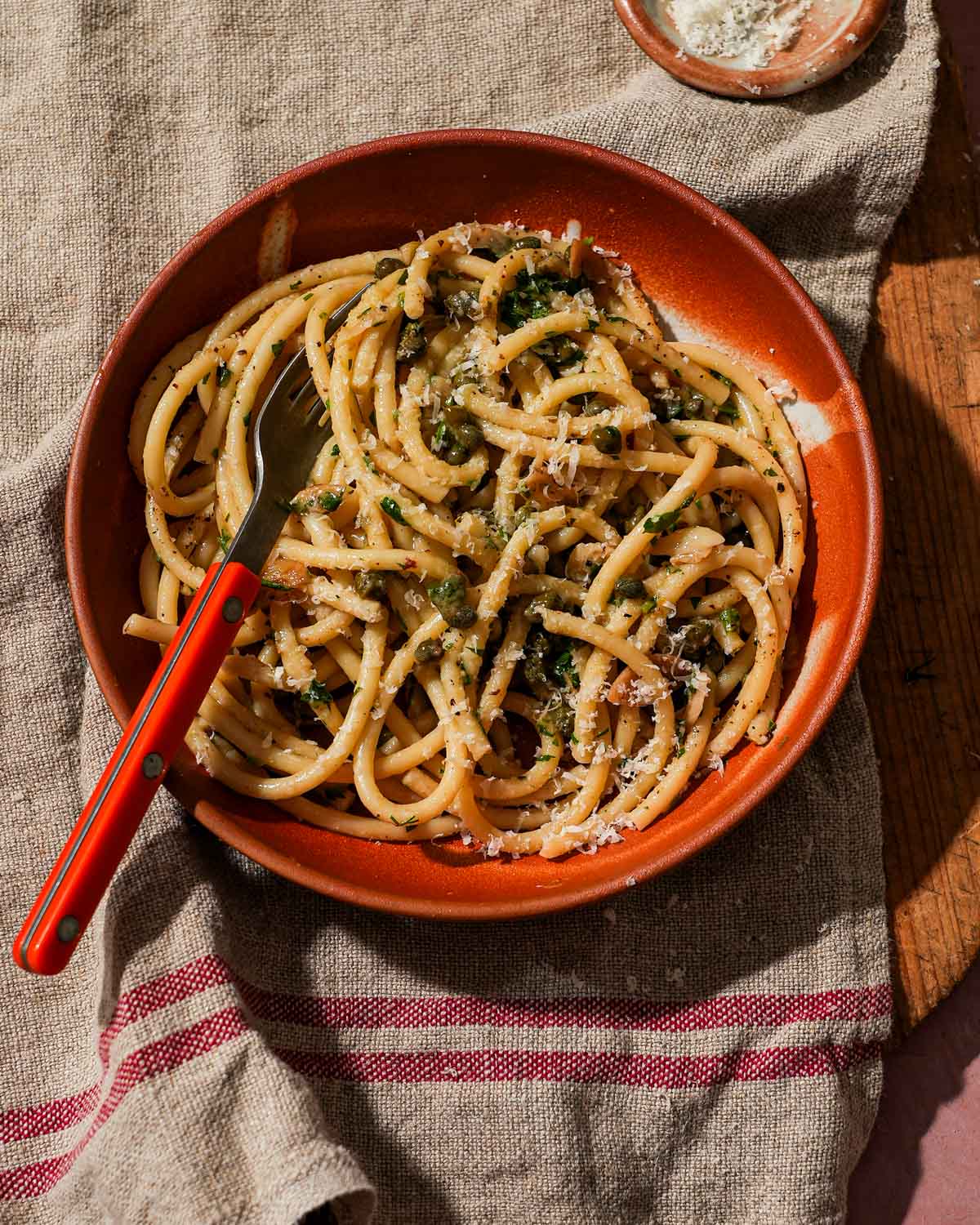 Pasta with capers and grated cheese in a red bowl, garnished with herbs.