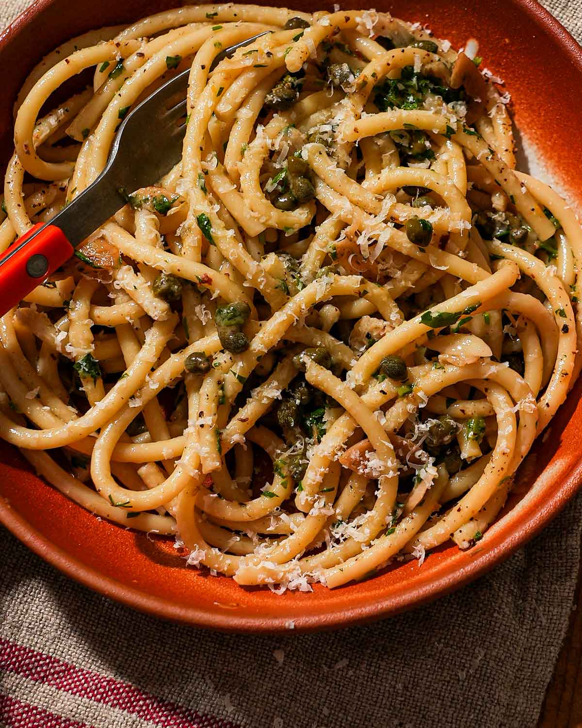 A fork twirls pasta with capers, cheese, and herbs in a red bowl.