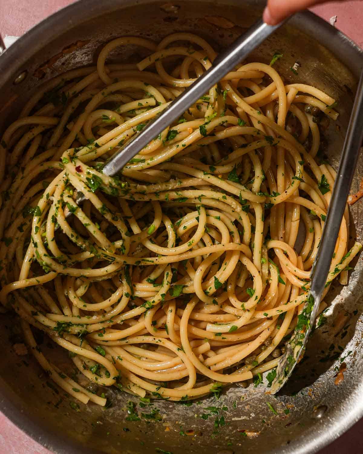 Stirring pasta with capers and herbs in a stainless steel pan.