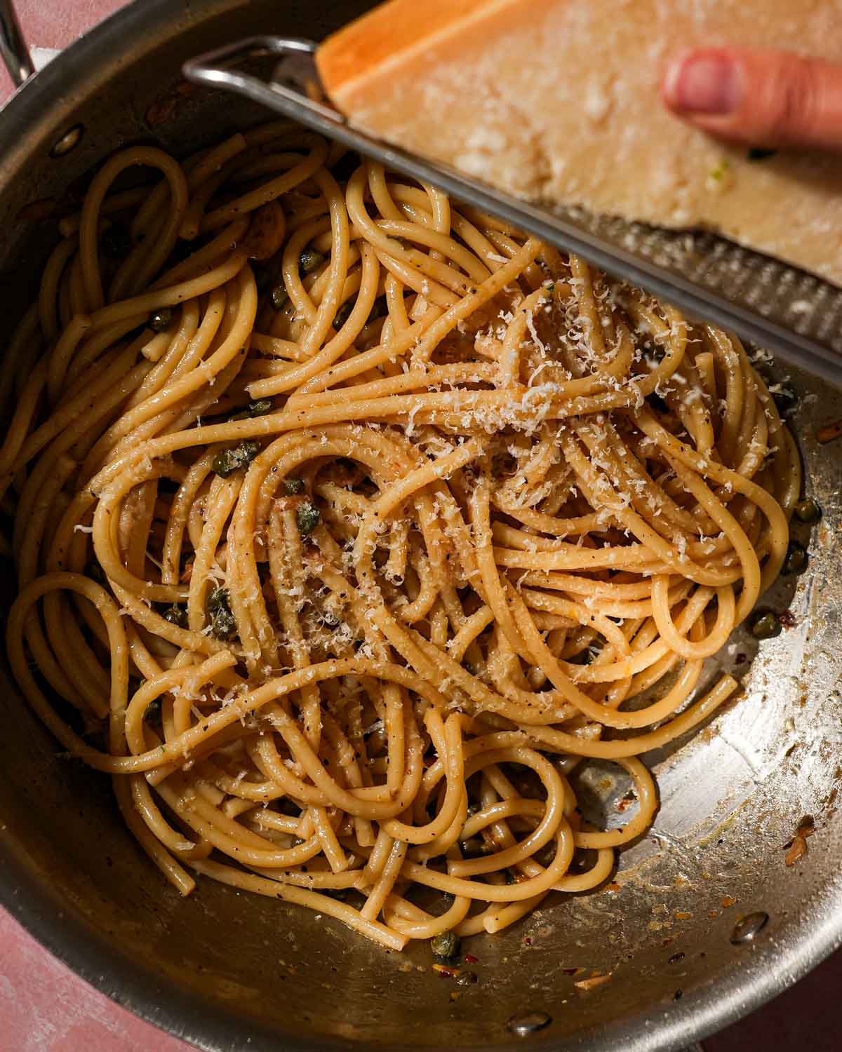 Grating Parmesan cheese over pasta with capers and herbs in a pan.