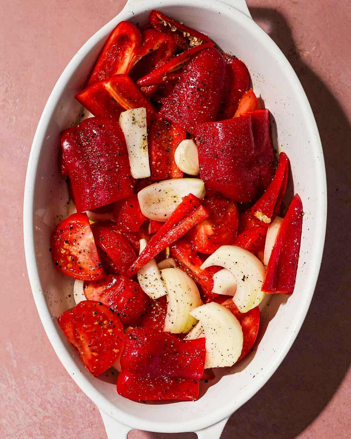 A white tray of red peppers, tomato, garlic, and onion ready to be roasted.