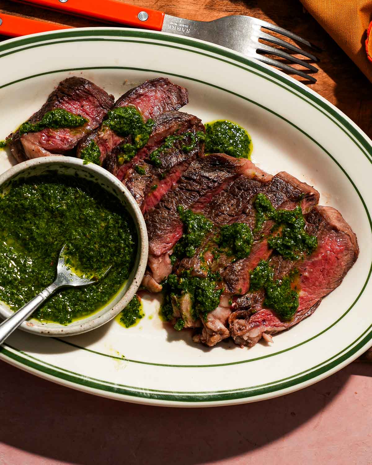 A plate of sliced ribeye steak with caper chimichurri sauce in a small bowl with a spoon.