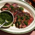 A plate of sliced ribeye steak with caper chimichurri sauce poured over and in a small bowl with a spoon.