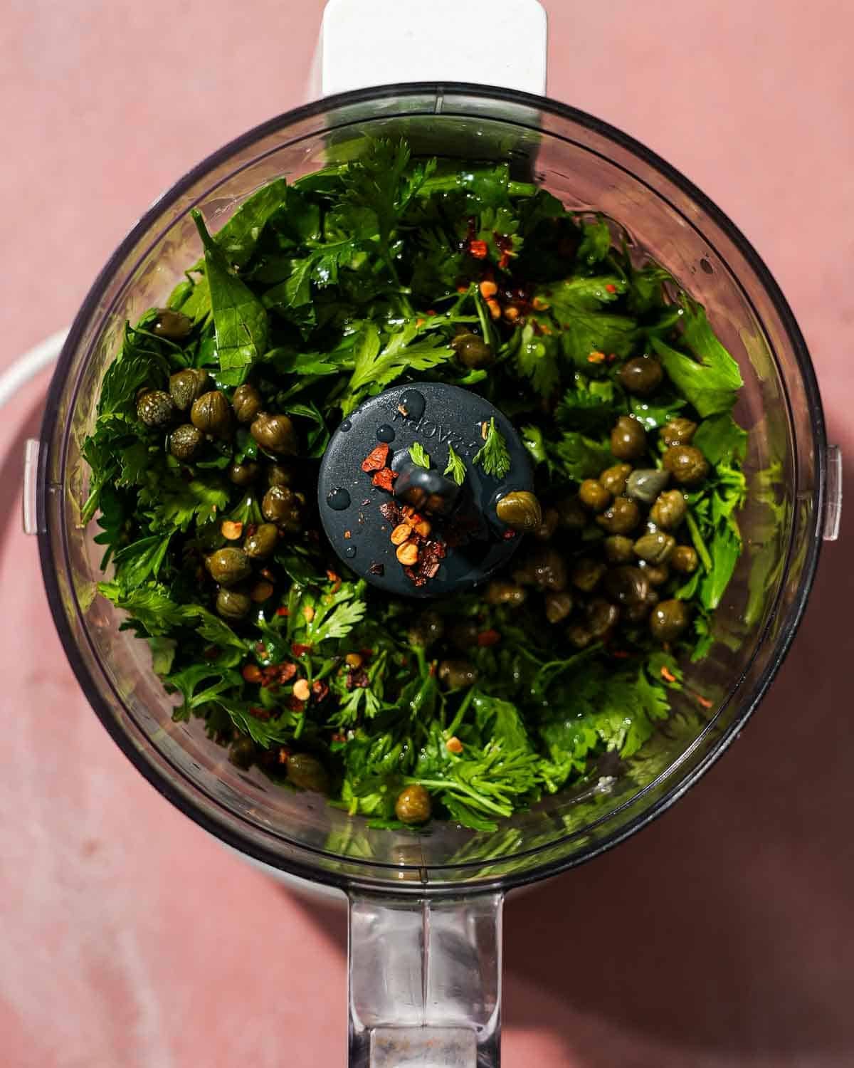 A food processor with all the ingredients for caper chimichurri sauce about to be blended.
